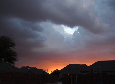 [A huge, dark, thick cloud covers the sky above the homes. There is a colorful swatch of yellow-orange which outlines the top of the buildings and one clear plastic basketball backboard. There is a hole in the cloud cover and through it one can see light blue skies with white clouds which must be at a distance beyond the storm.]